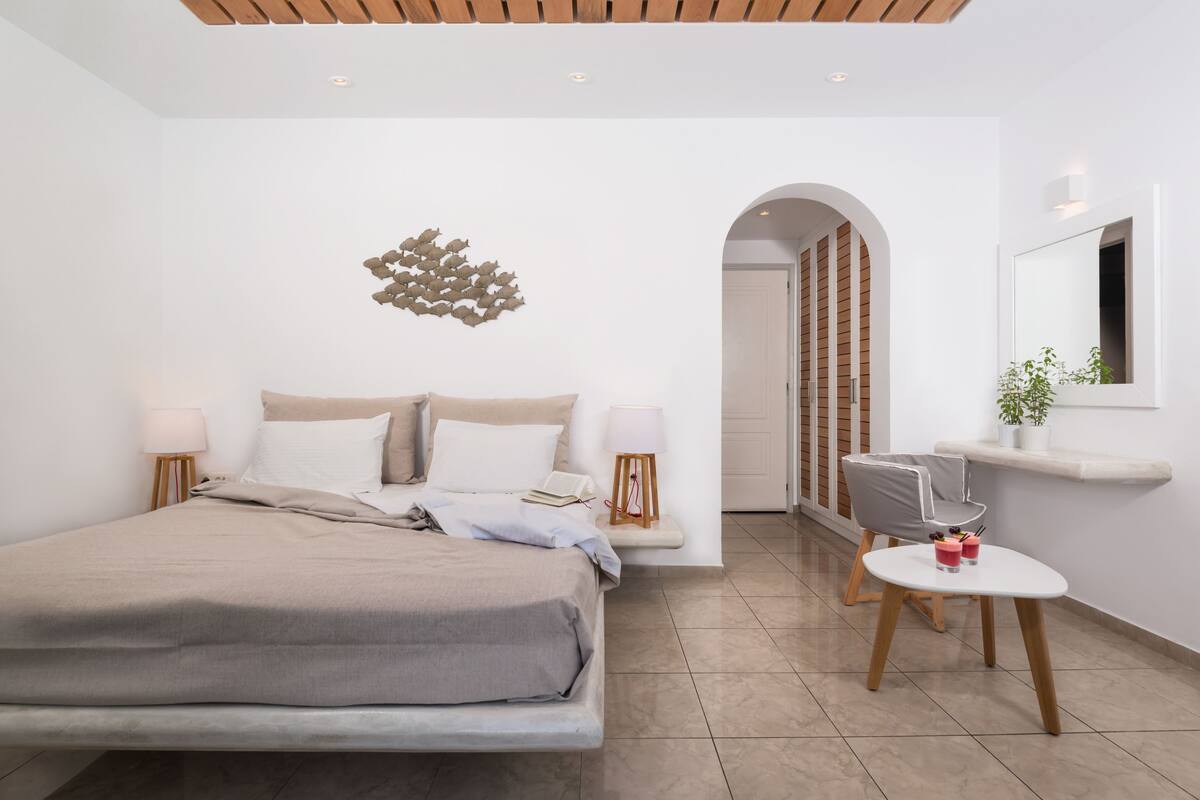 A modern bedroom with a large bed, bedside lamps, a small table with drinks, and a mirror on the wall.