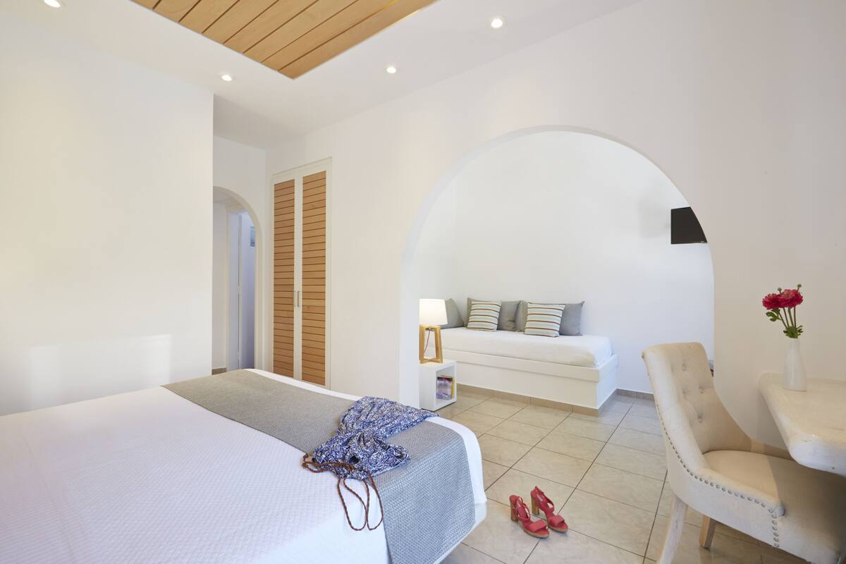 A modern hotel room with a large bed, a seating area with a sofa and armchair, a small table with a lamp, and a decorative vase with flowers.