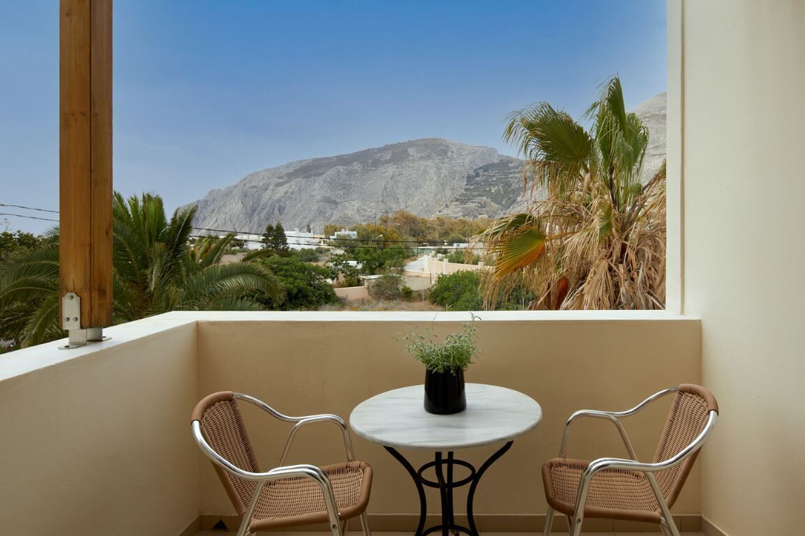 A balcony with two wicker chairs and a small round table, overlooking a mountainous landscape with sparse vegetation and buildings.