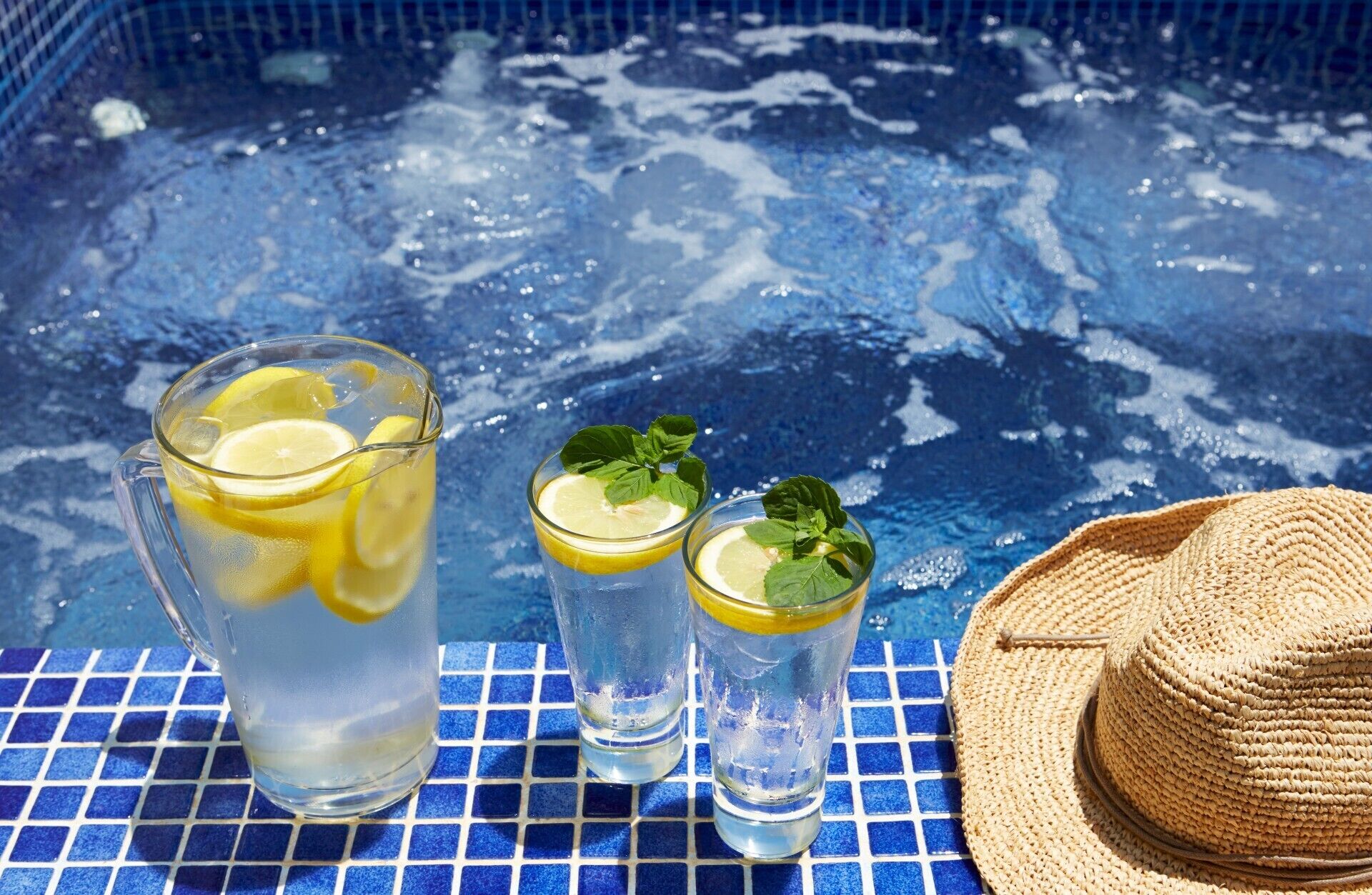 A glass pitcher and two glasses of lemonade with lemon slices and mint leaves, placed on a blue tiled surface near a pool.