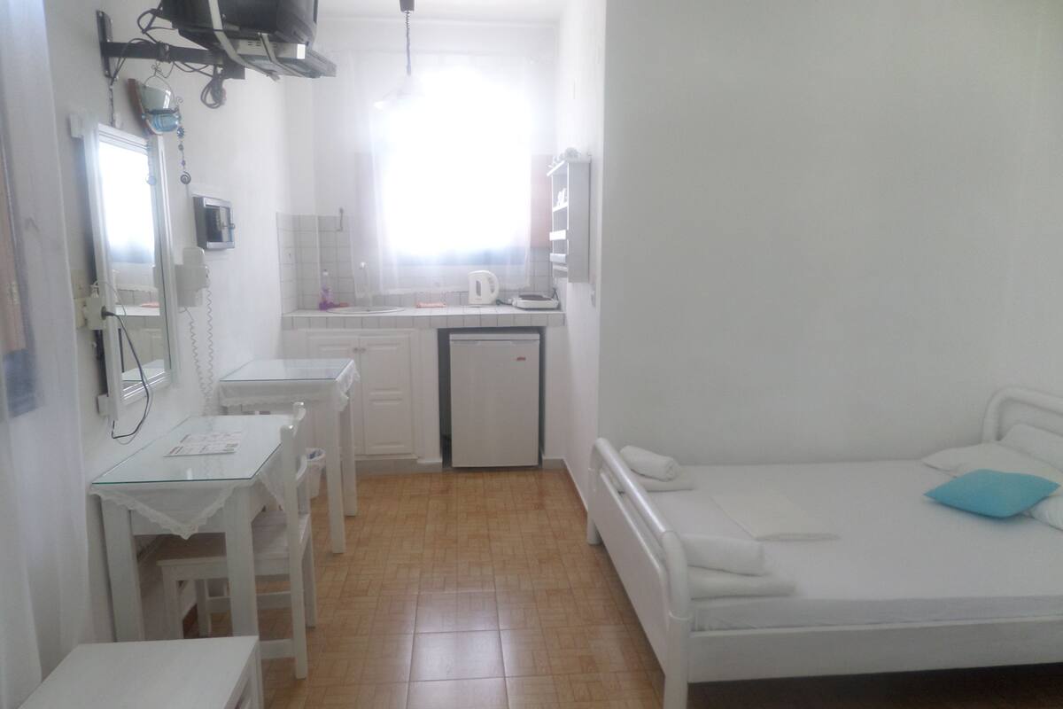 A small, white room with a bed, a desk, and a small kitchenette.