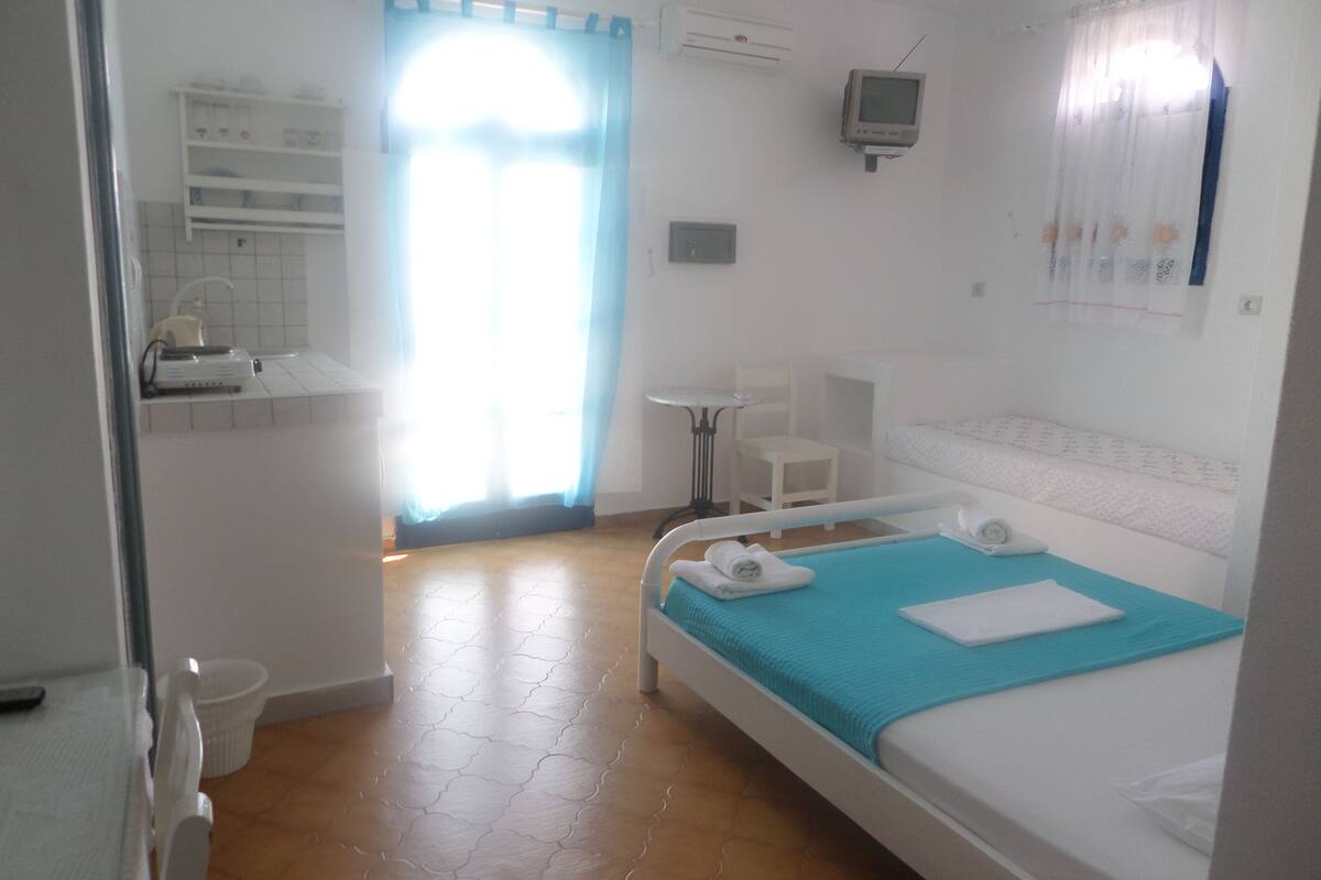 A bedroom with a bed, a small table, a chair, a TV, and a bathroom visible through an open door.