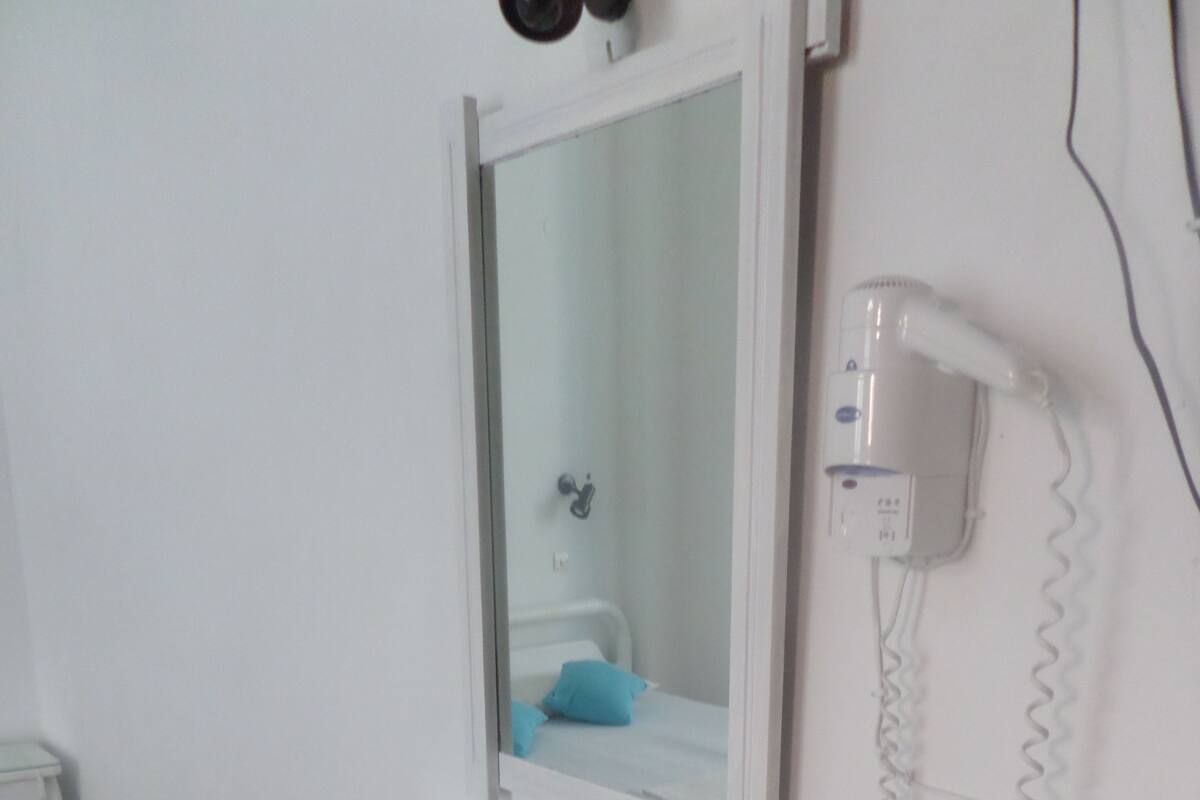 A small, white room with a glass-top table and a chair, a mirror above the table, and a hairdryer on the wall.