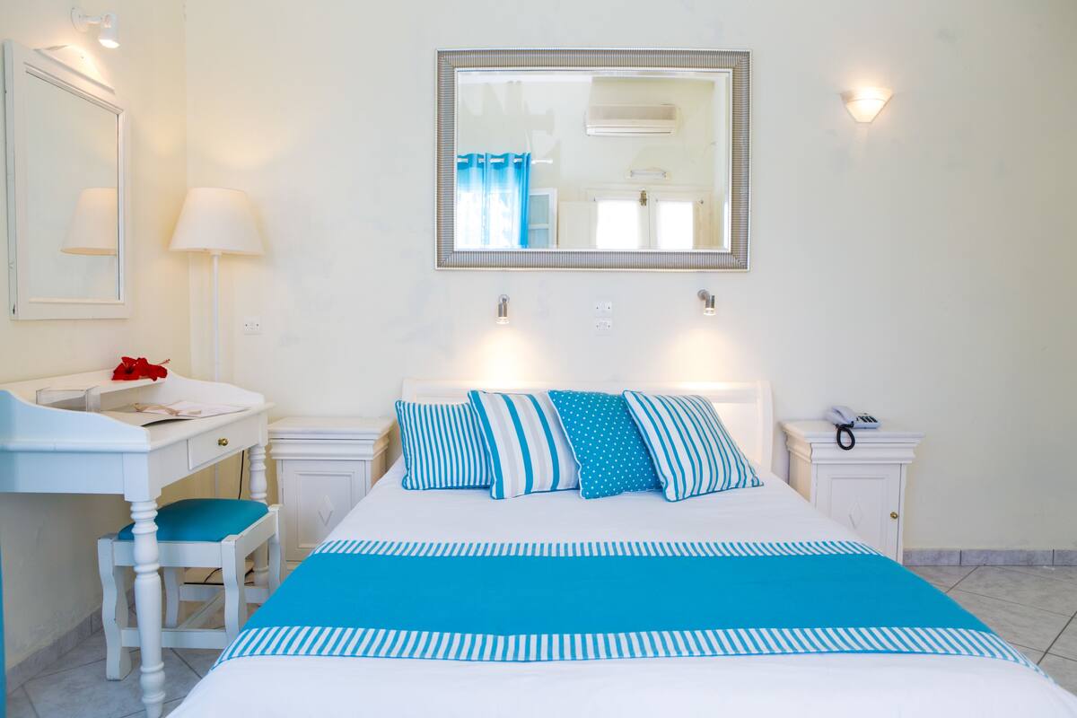 A bedroom with a bed, a desk, a chair, a nightstand, a mirror, and a wall-mounted lamp.