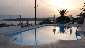 Una piscina con solárium de piedra, rodeada de sillas de descanso y sombrillas, con vistas al mar y colinas al fondo.