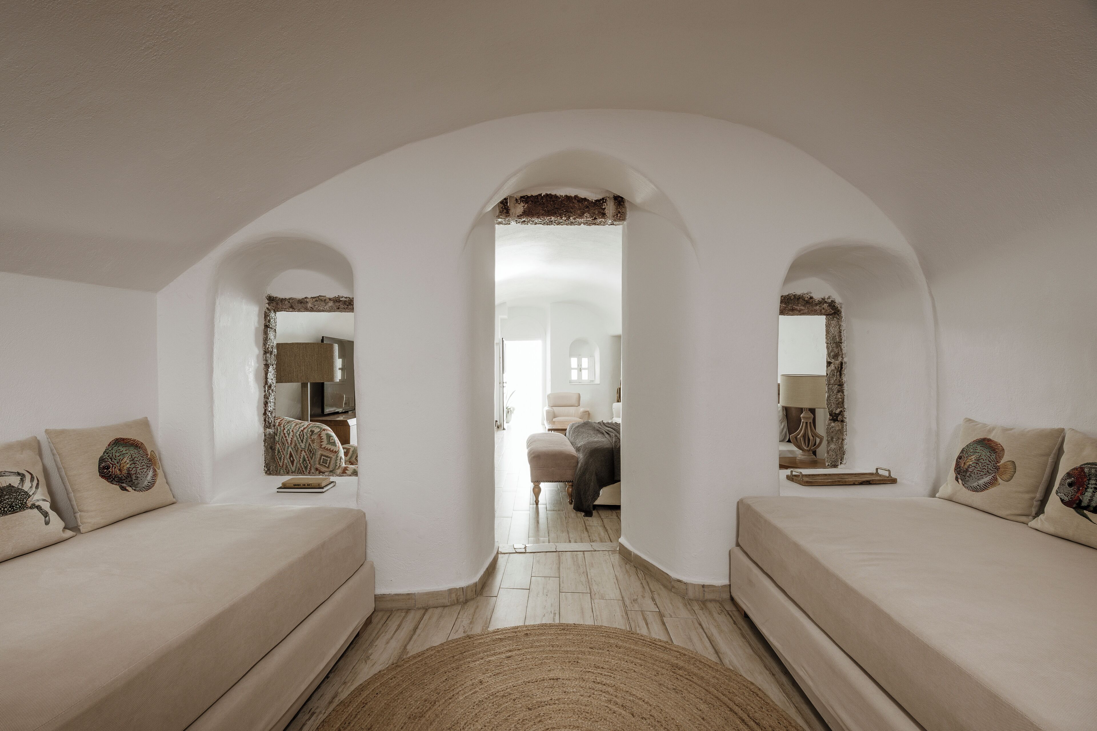 A modern hotel room with a minimalist design, featuring two sofas with decorative pillows, a central archway leading to another room, and a wooden floor.