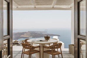 A round table with four wicker chairs, a book, and a decorative bowl on a balcony overlooking a scenic coastal view. - Aqua Luxury Suites (Santorini)
