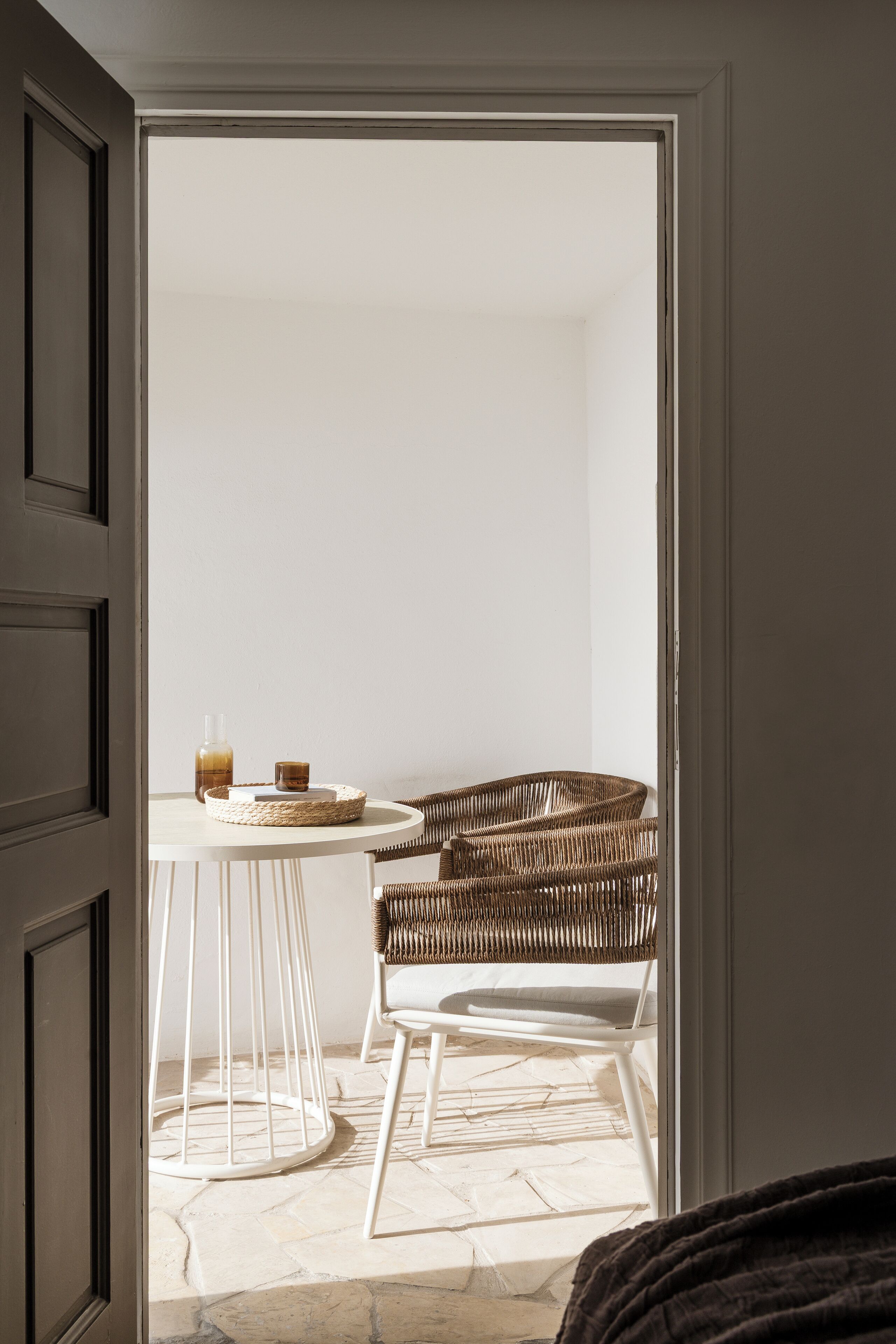 An open door leading to a room with a wicker chair, a small round table, and a bottle.