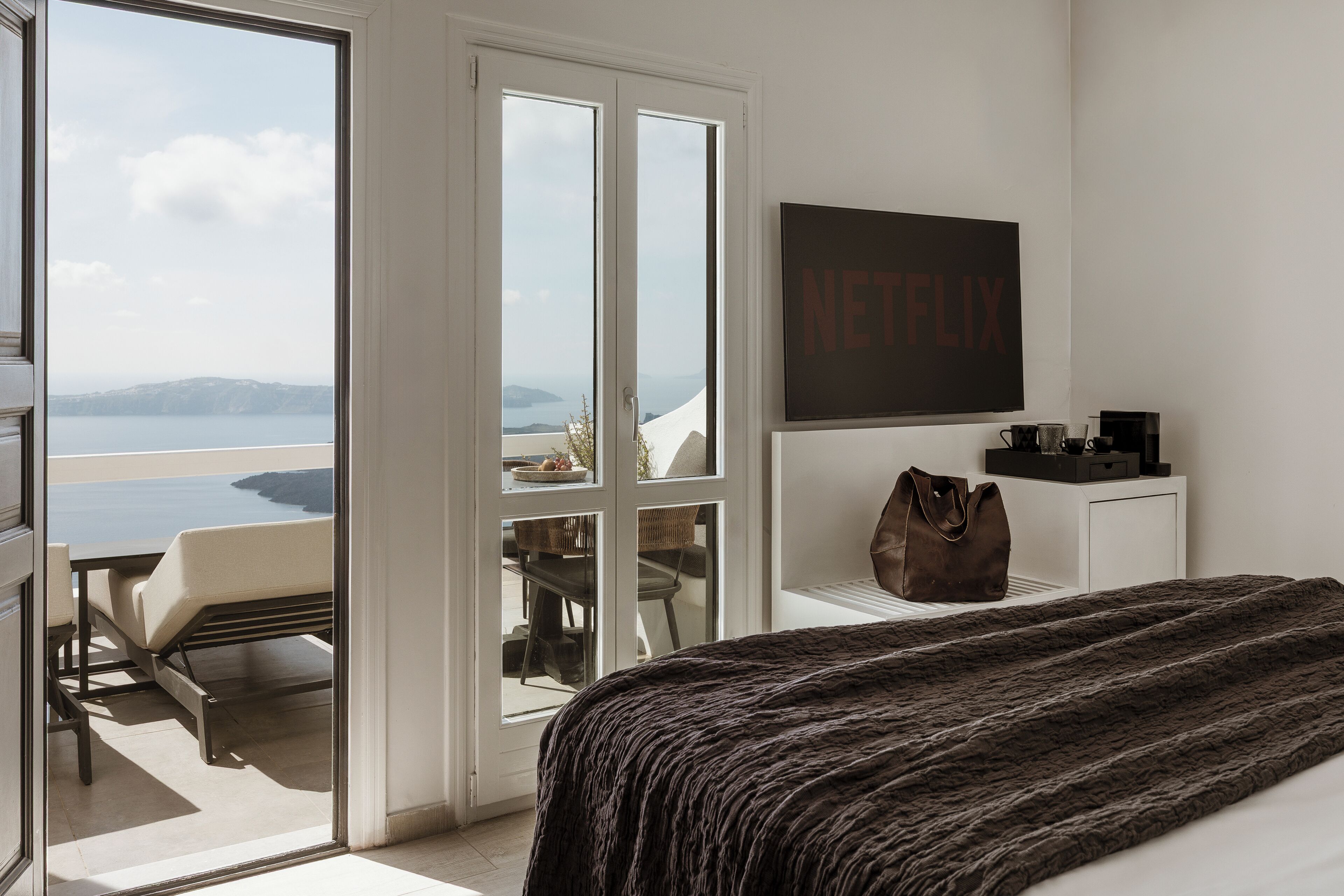 A bedroom with a large bed, a view of the sea through a sliding door, a Netflix sign on the wall, and a small table with a bag.