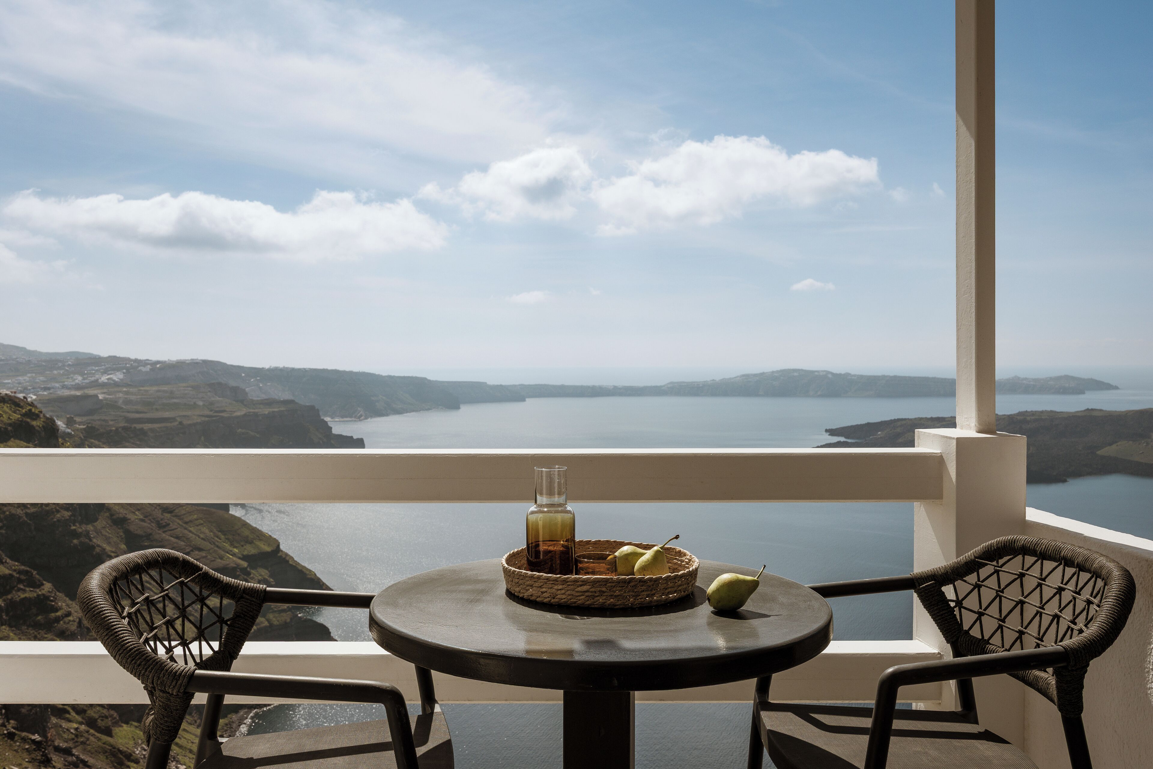 A balcony with wicker chairs, a table with a basket of fruit and a bottle, and a view of a coastal landscape.