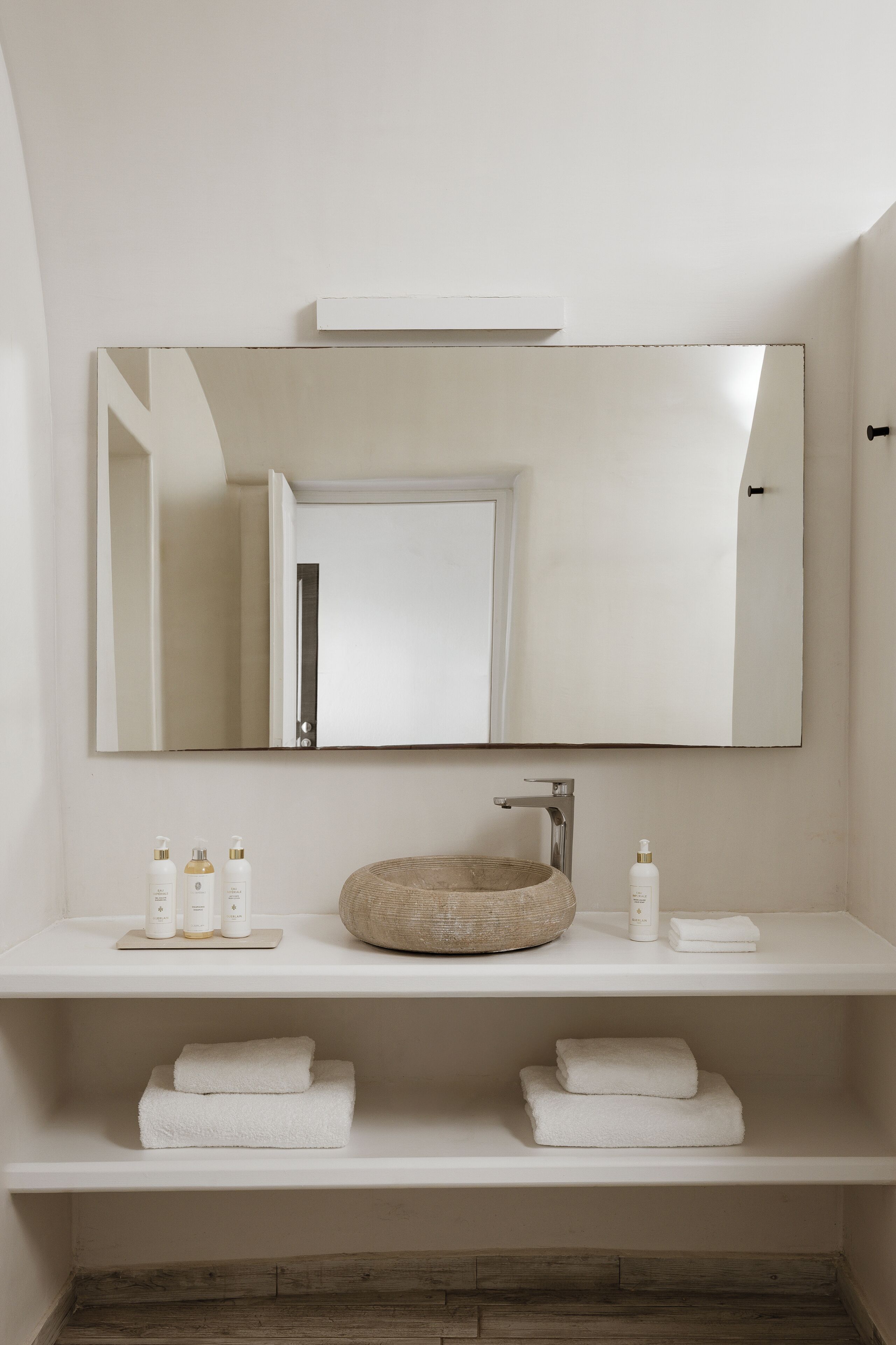 A bathroom with a white vanity, a round sink, and a mirror above it. There are two shelves with towels and toiletries.