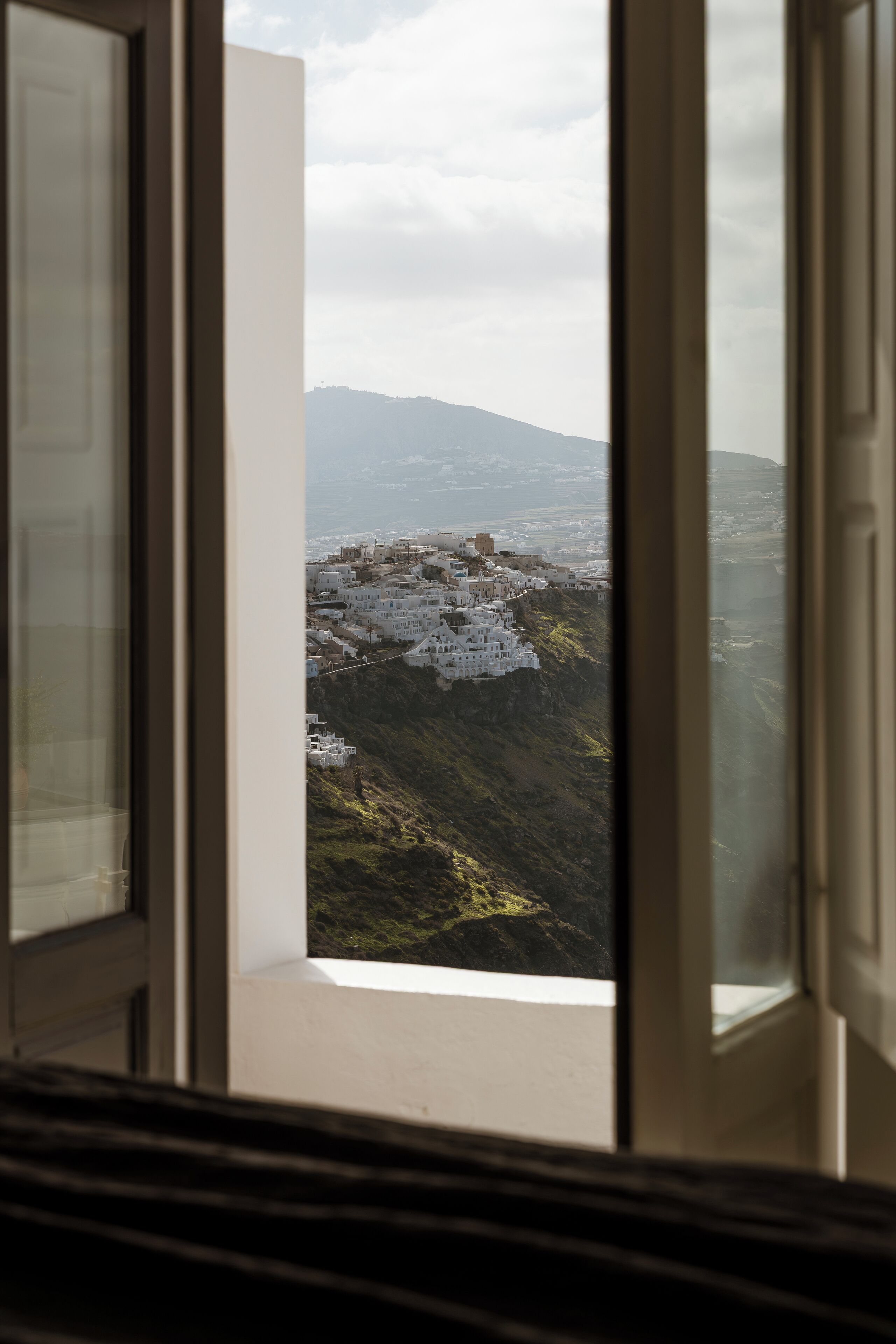 Vista a través de una ventana de un pueblo costero con edificios blancos, una montaña al fondo y cielo despejado.