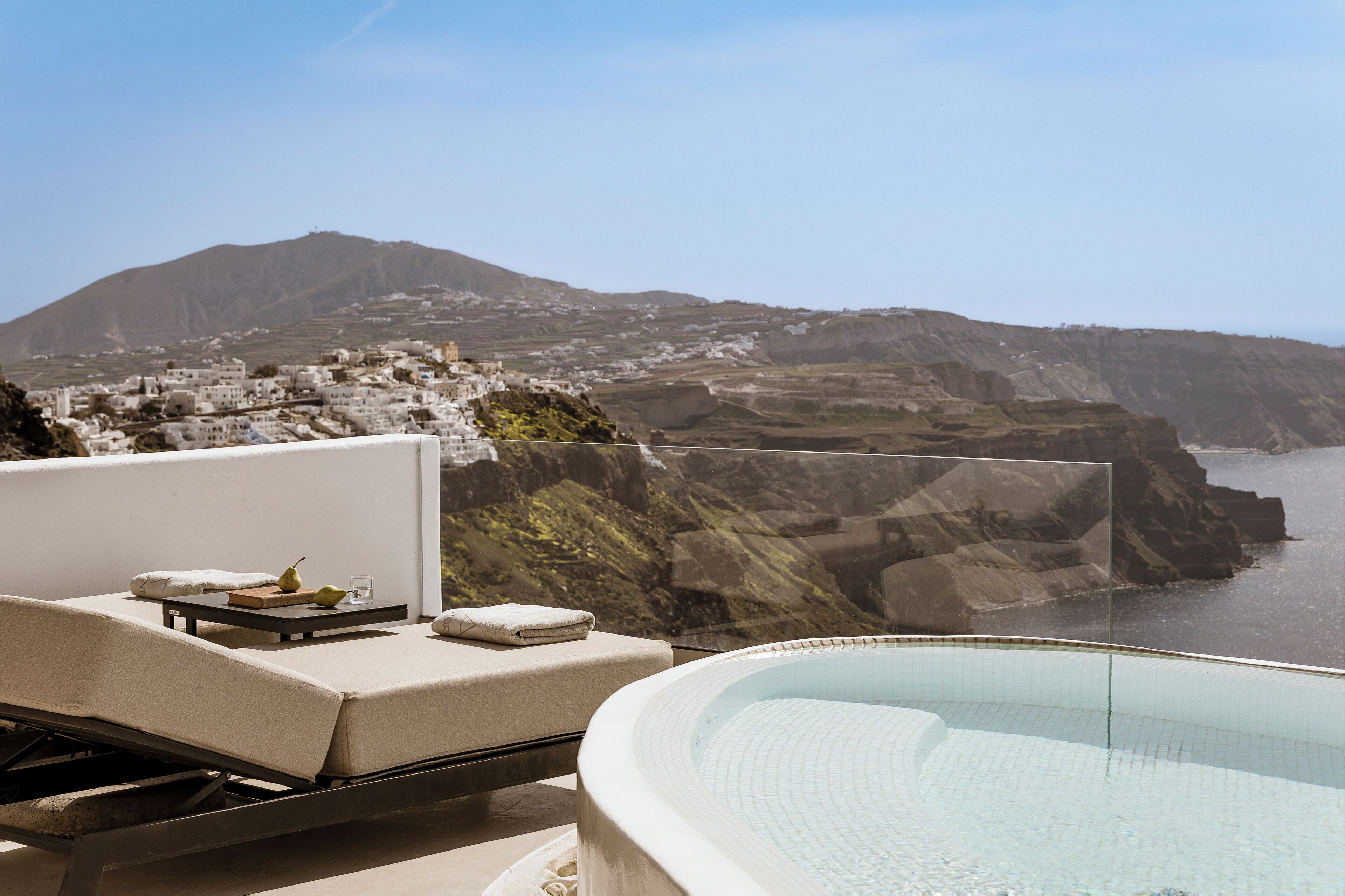 A sunlit outdoor lounge area with a pool, a view of a coastal town, and a mountain in the background.