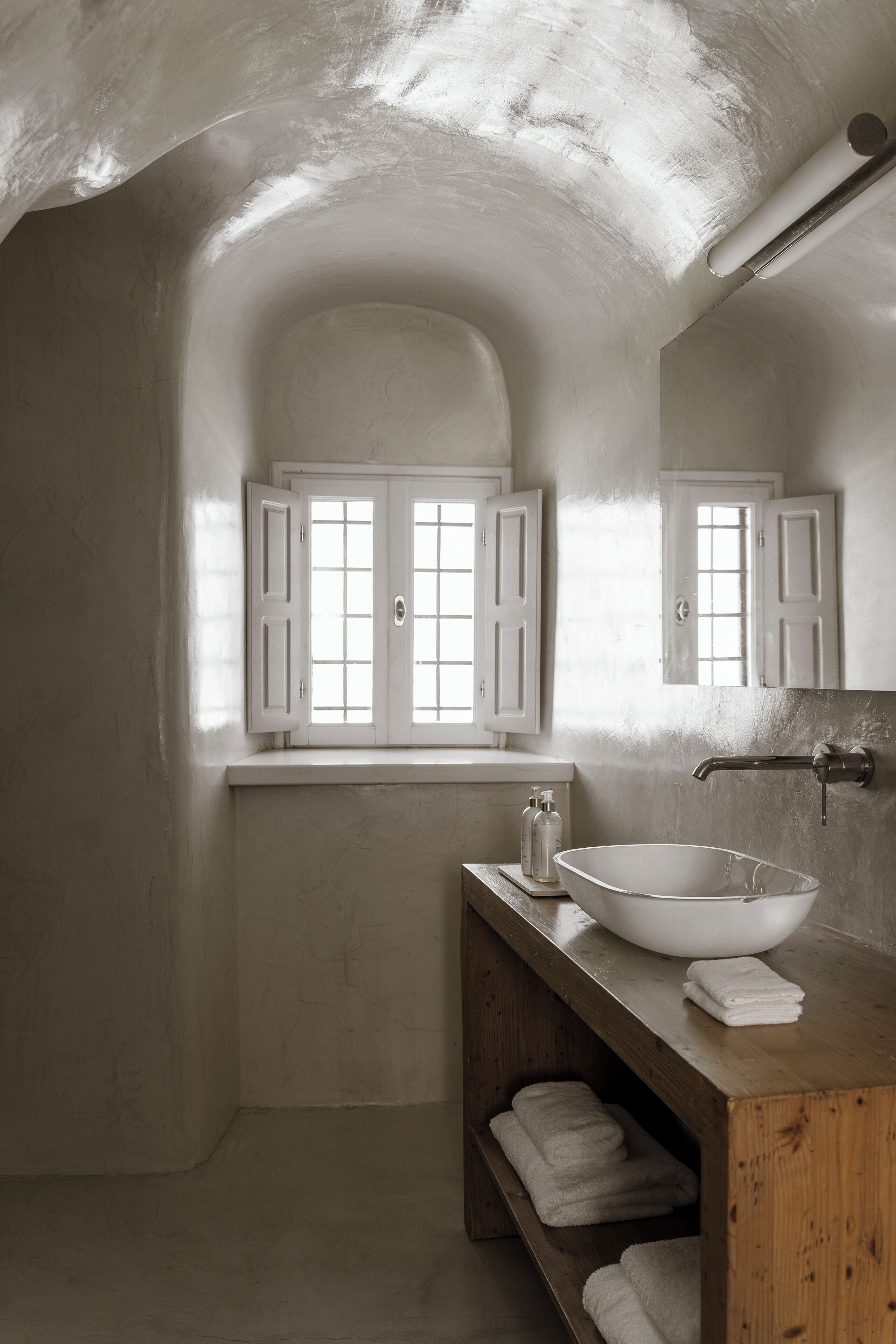 Une salle de bain avec un grand lavabo blanc, un meuble-lavabo en bois et une fenêtre dont les volets sont ouverts.