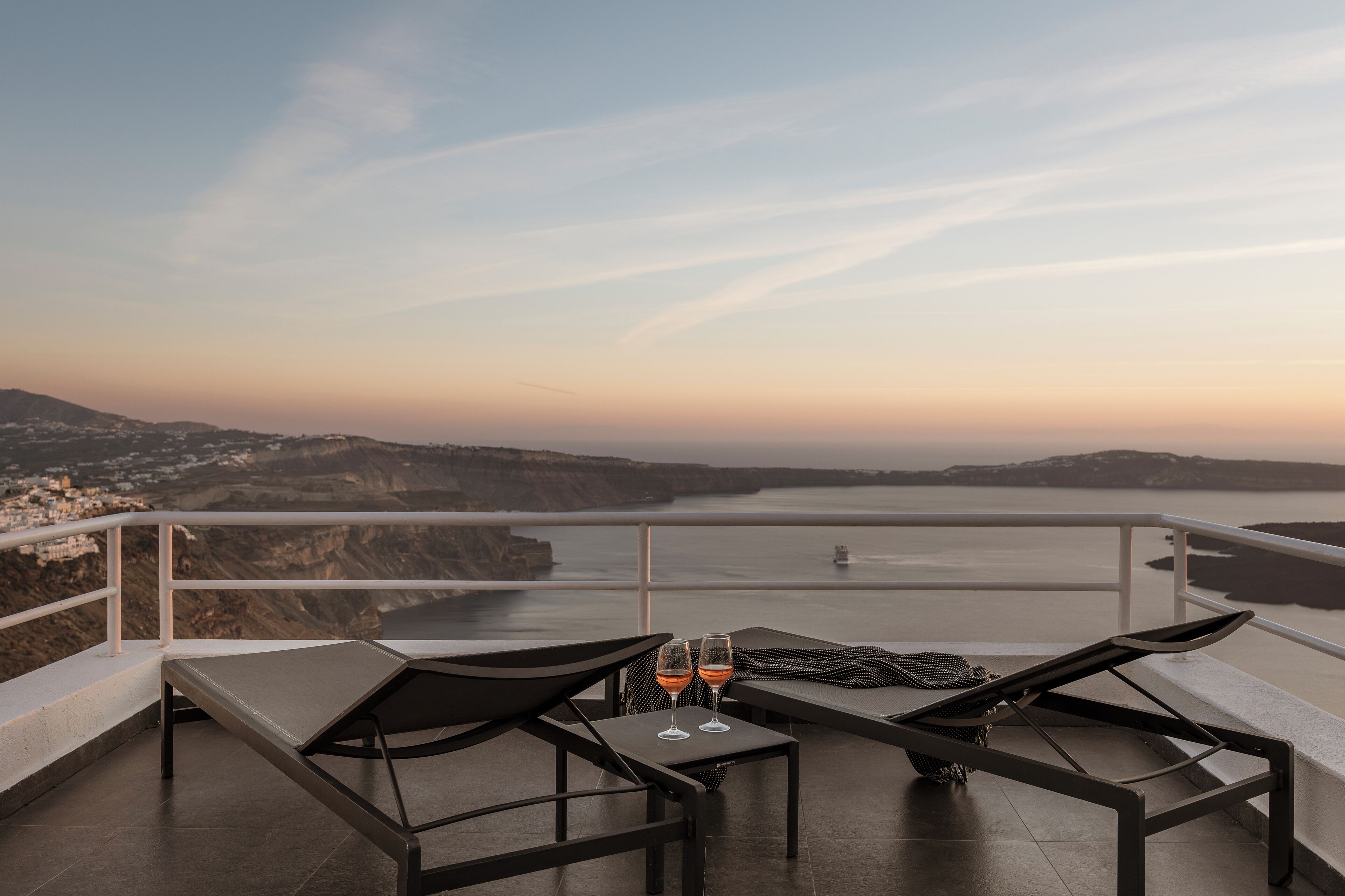 A terrace with two lounge chairs, a table, and two glasses of wine, overlooking a coastal landscape with cliffs and a bridge.