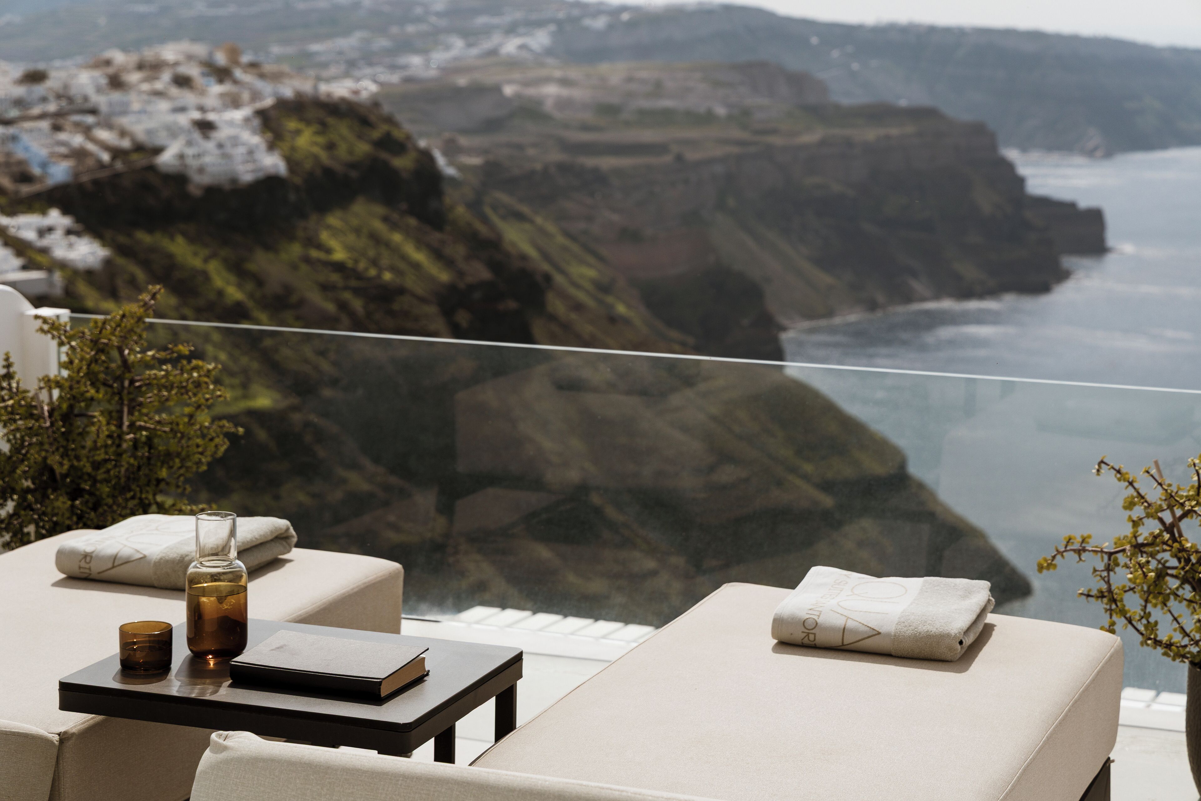 A balcony with a view of cliffs and the sea, a table with a book and a candle, and two lounge chairs with towels.