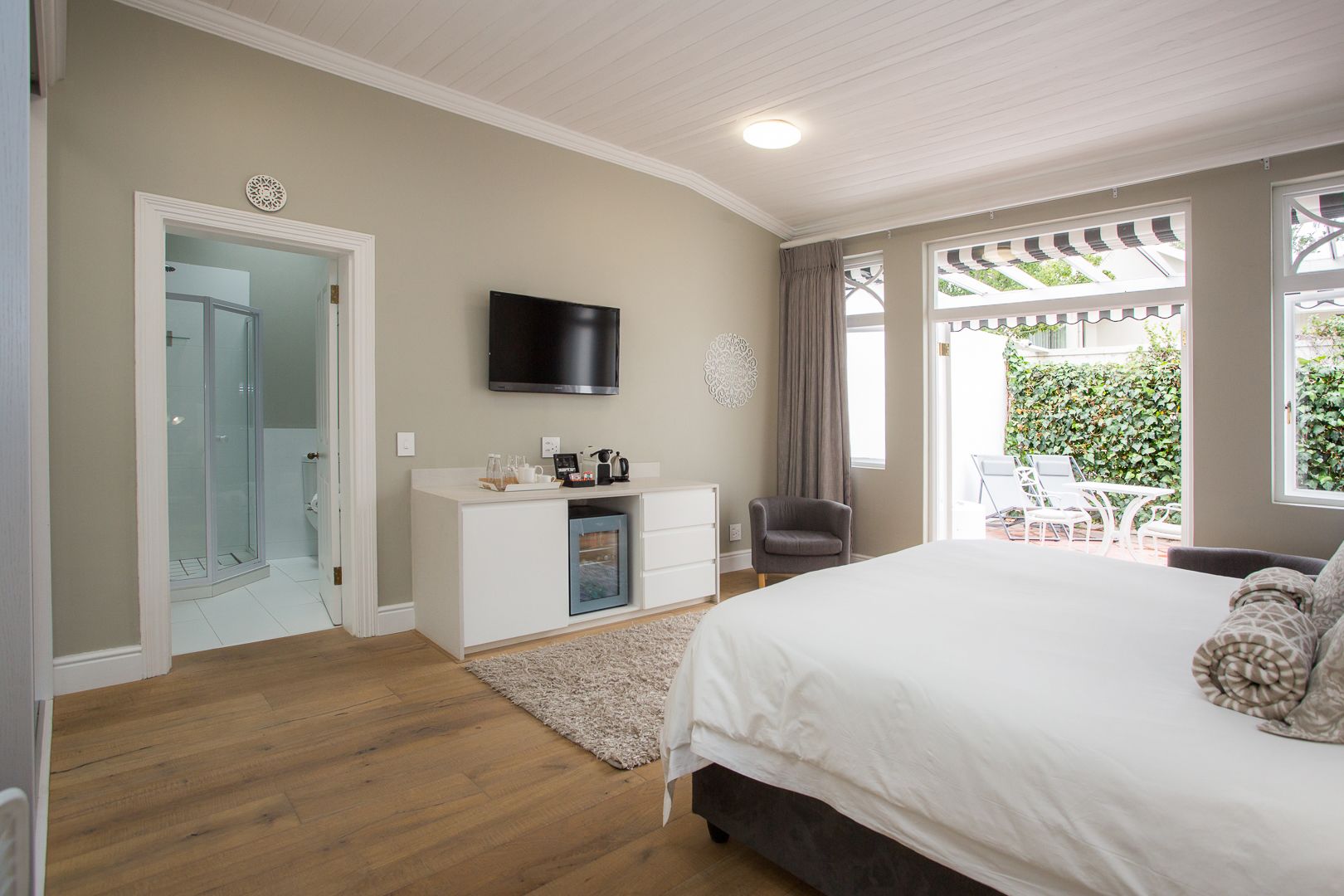 A bedroom with a large bed, a TV mounted on the wall, a desk with a computer, a chair, and a view of a garden through the window.