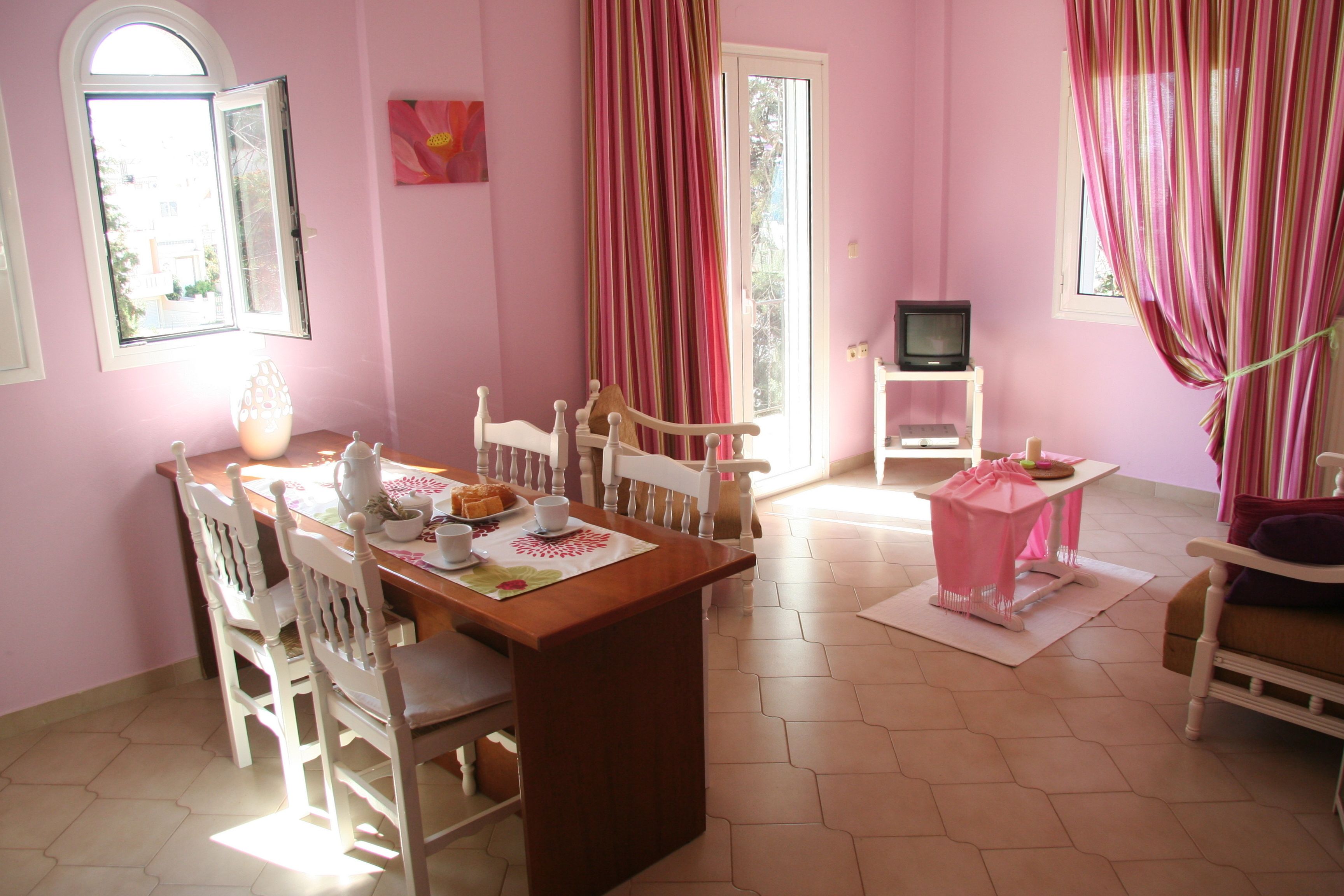 A dining area with a wooden table and white chairs, a television on a stand, and a pink curtain.