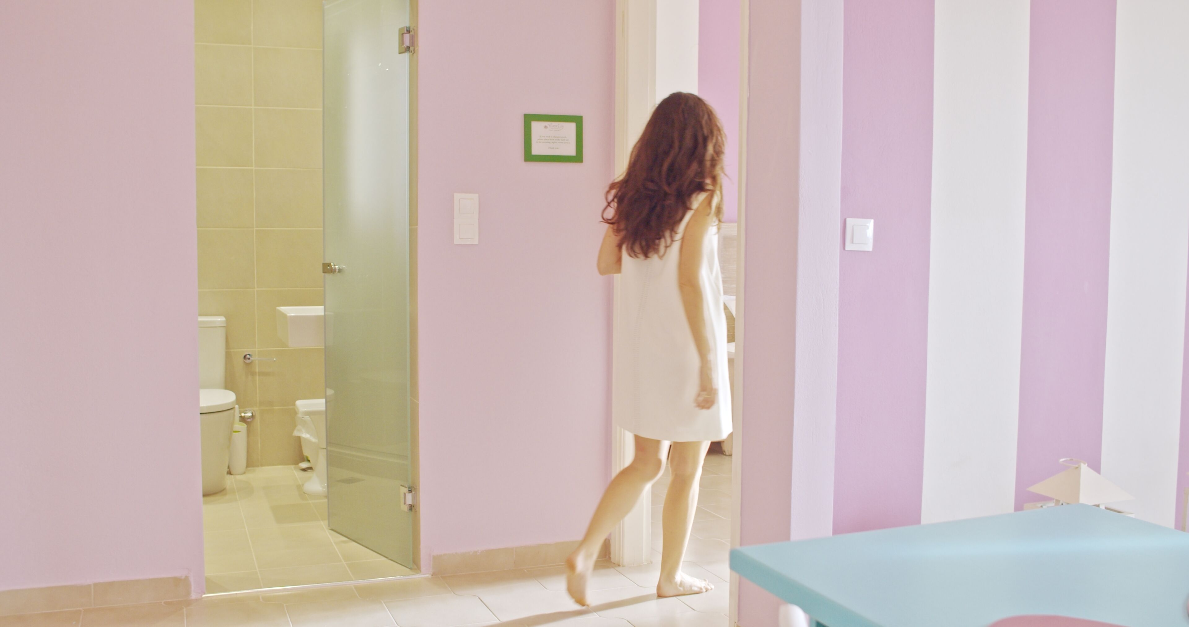 Een vrouw in een badjas loopt door een badkamer met een glazen douchecabine, een wastafel en een toilet.