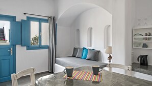 A room with a bed, a table with two mugs, a window with curtains, and a white wall with a shelf.