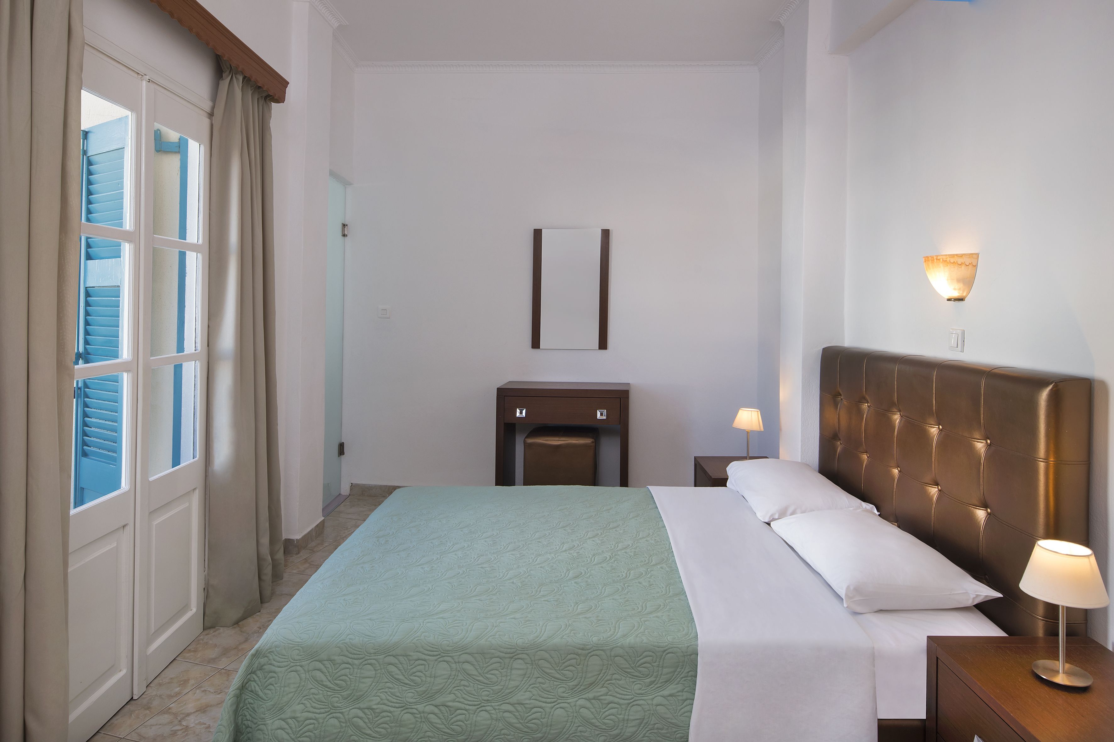 Quarto de hotel com cama, mesa de cabeceira, luminária na parede e porta com cortinas.