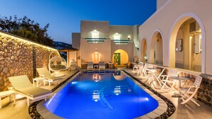 A swimming pool with lounge chairs and tables, surrounded by a stone wall and a building with arches.