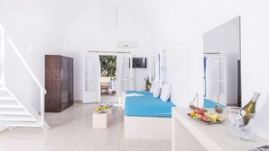 A bright, white room with a bed, a wooden cabinet, a small table with fruit and a bottle, and a large mirror. - Bella Santorini (Santorini)