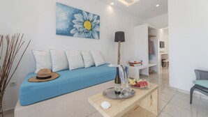 A modern living room with a blue sofa, a wooden coffee table with a bottle and glasses, and a painting of a flower on the wall. - Bella Santorini (Santorini)