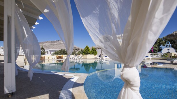 A clear blue pool with white curtains, a white building, and a mountain in the background. - Bella Santorini (Santorini)