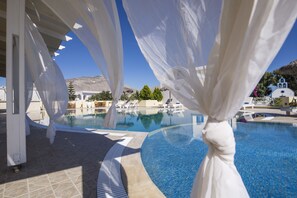 A clear blue pool with white curtains, a white building, and a mountain in the background. - Bella Santorini (Santorini)