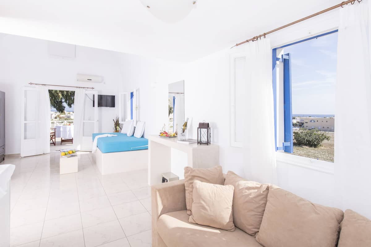 A bright, white room with a bed, a sofa, and a TV. There are windows with blue frames and a view of the outside.