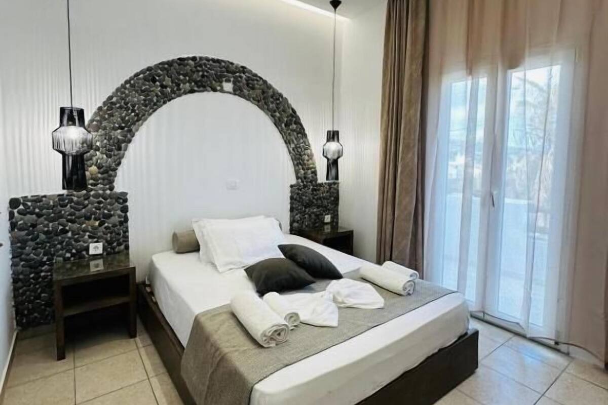 A modern bedroom with a stone arch headboard, a large bed with white linens, two bedside tables, and a view of the outdoors through a window with curtains.