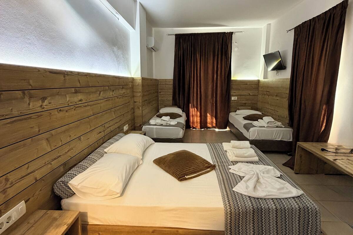 A hotel room with a large bed, two smaller beds, wooden headboard, a flat-screen TV, and a bench.