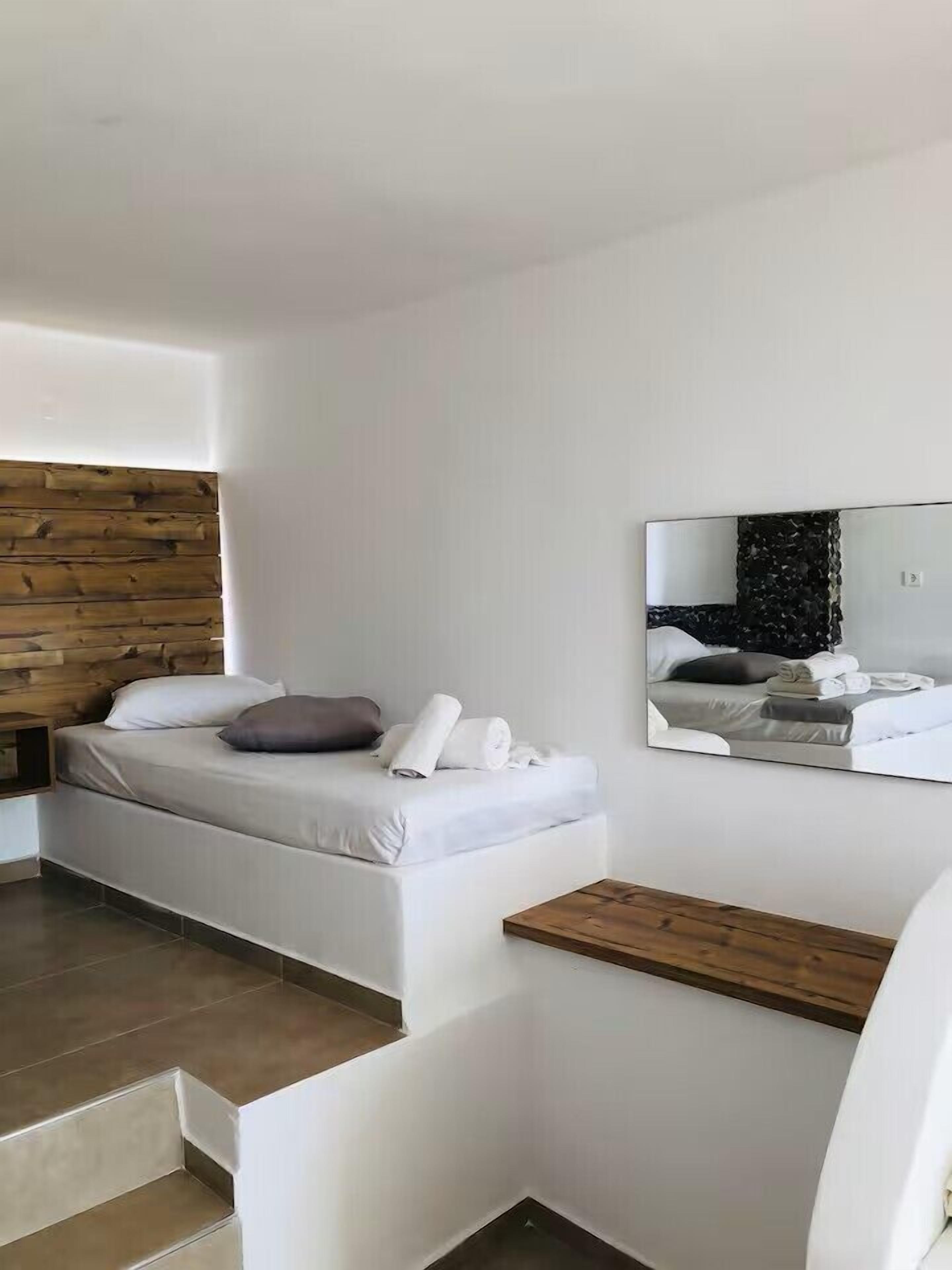 A modern bedroom with a wooden headboard, a bed with white linens and a rolled towel, a wooden nightstand, and a large mirror on the wall.