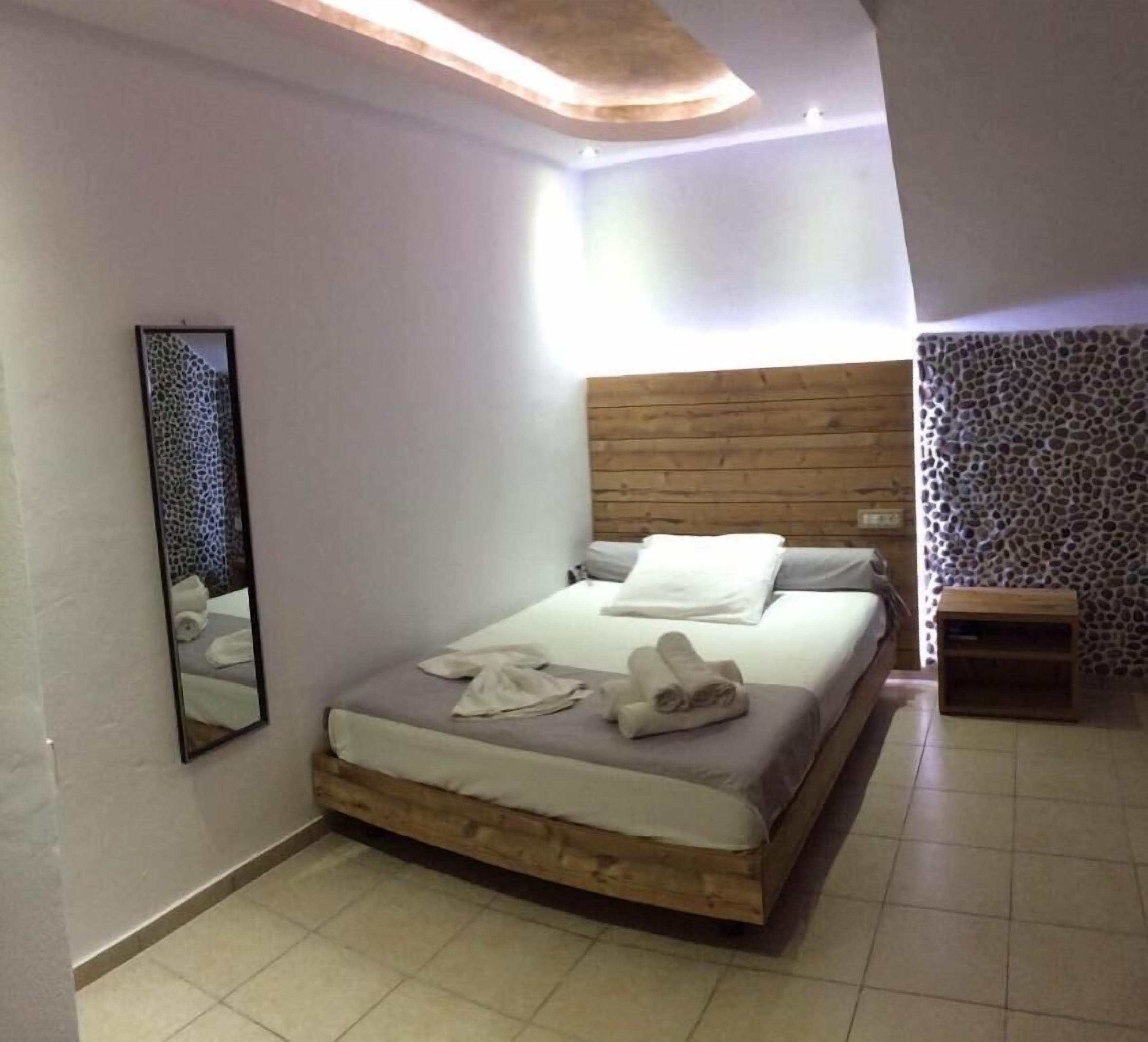 Un dormitorio con cabecera de madera, cama con ropa blanca, mesita de noche de madera, un espejo y una pared con diseño.