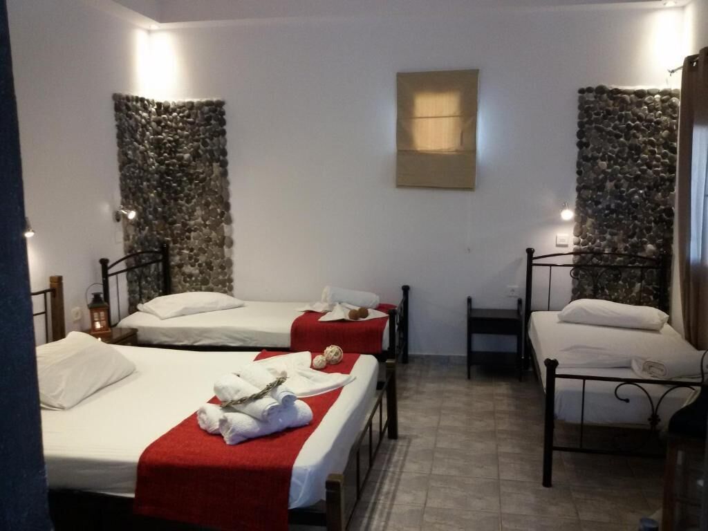 A room with three beds, each with a white mattress and red bedspread. There is a small table with a chair, a wall-mounted light, and a stone wall decoration.