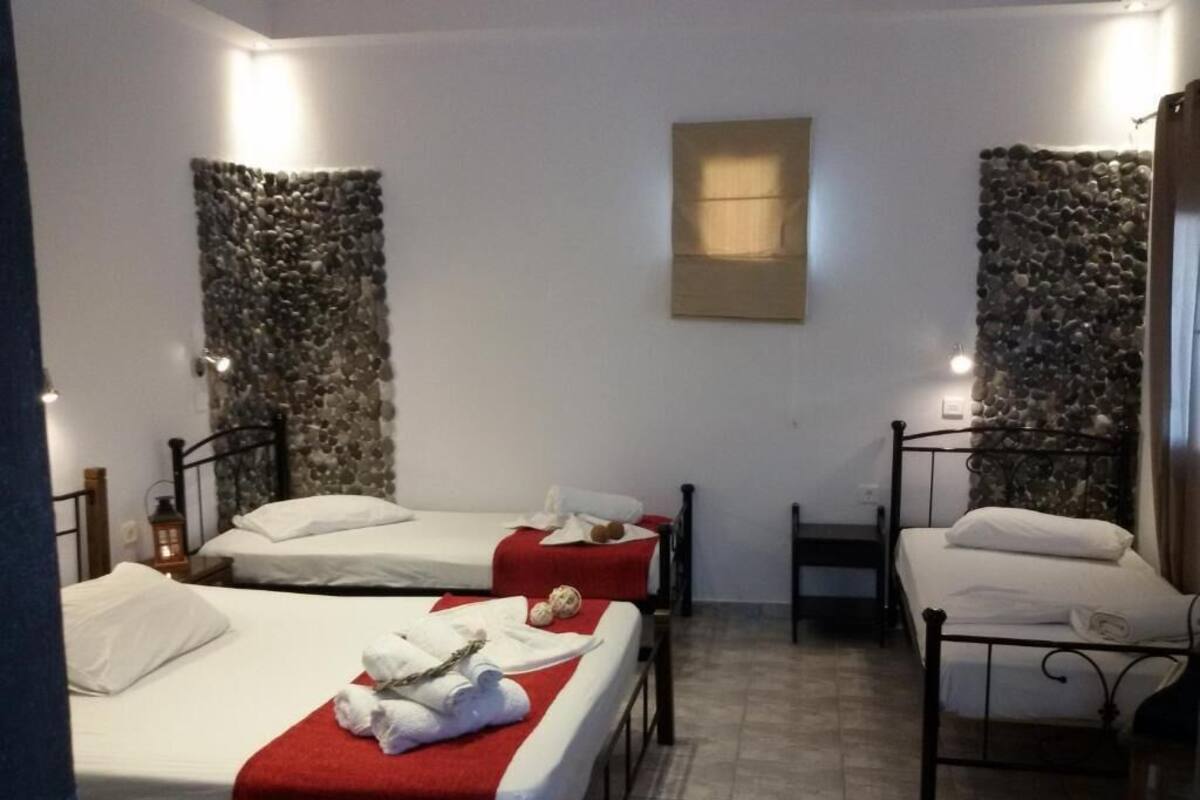 A room with three beds, each with a white mattress and red bedspread. There is a small table with a chair, a wall-mounted light, and a stone wall decoration.