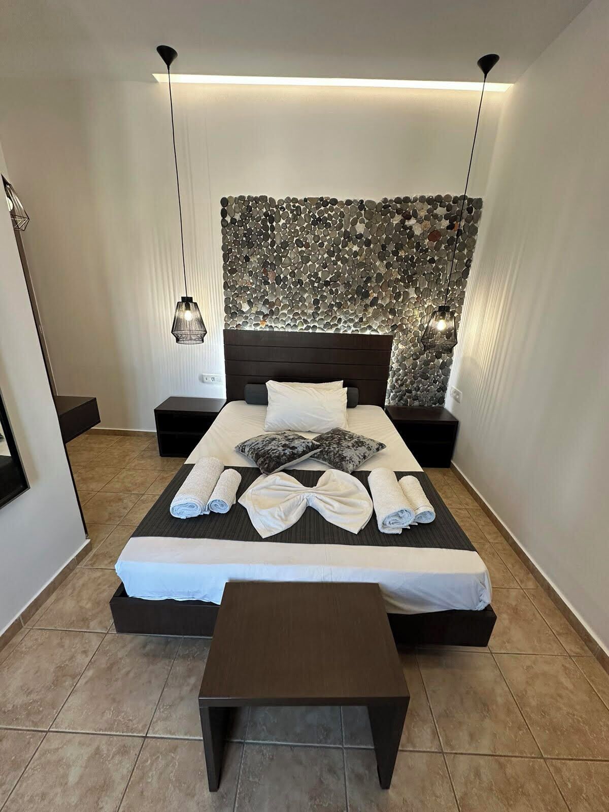 A modern bedroom with a stone wall headboard, a neatly made bed with towels, and a small table.