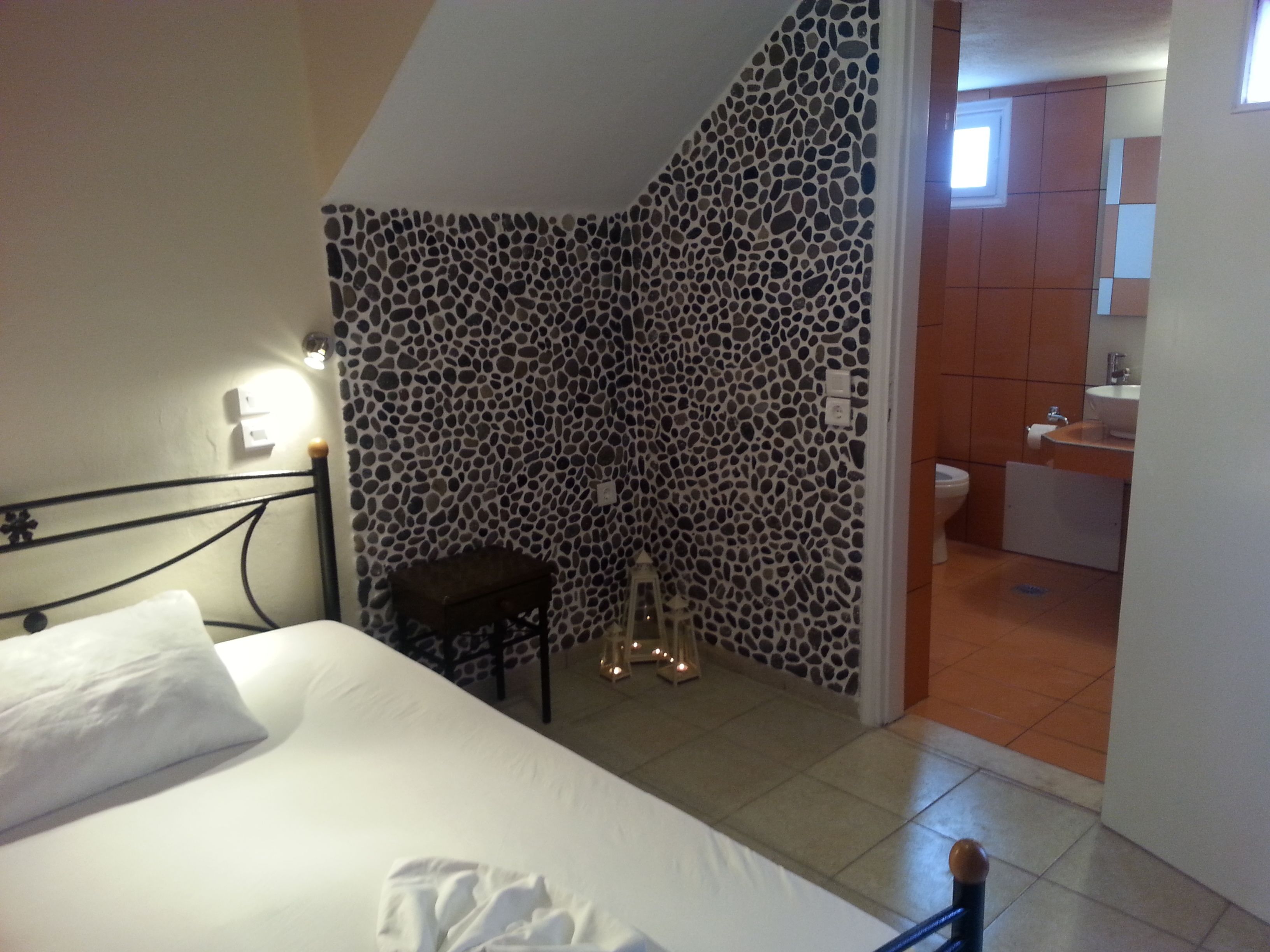 A bedroom with a bed, a nightstand, a wall with a mosaic pattern, a small stool, and a doorway leading to a bathroom.