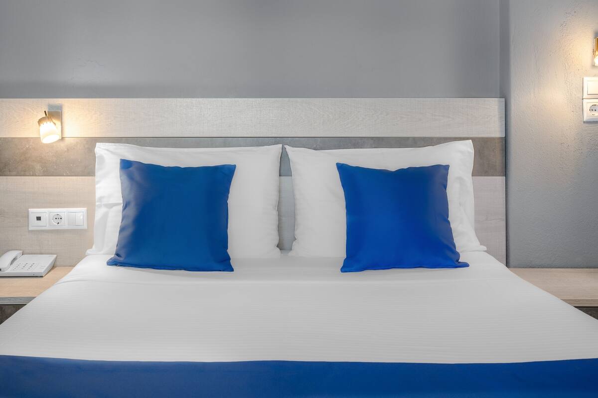 A hotel room with a large bed, two blue pillows, a bedside phone, and wall-mounted light fixtures.