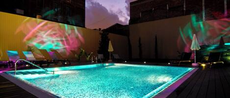 An outdoor swimming pool with a wooden deck, lounge chairs, and a projection of abstract shapes on the wall.