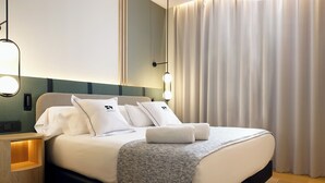 A neatly made bed with white linens and pillows, a bedside table with a lamp, and a large window with curtains. - Hotel & Spa Villa Olímpic@ Suites (Barcelona)