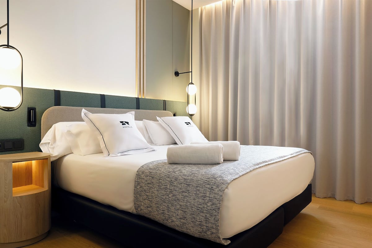 Ein ordentlich bezogenes Bett mit weißen Leinen und Kissen, ein Nachttisch mit Lampe sowie ein großes Fenster mit Vorhängen.