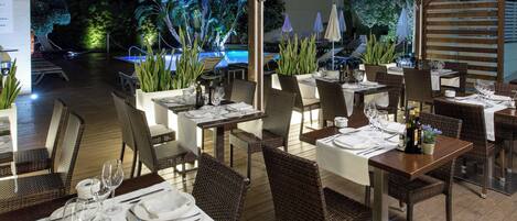 An outdoor dining area with tables and chairs, a pool in the background, and potted plants.
