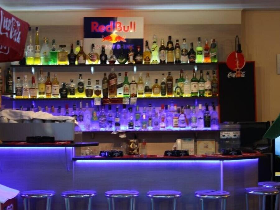 A bar area with multiple shelves of liquor bottles, a Red Bull sign, and a Coca-Cola refrigerator. There are four stools in front of the bar.
