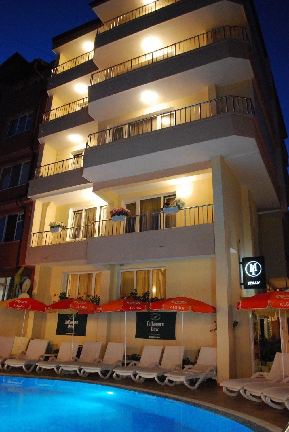 Un hôtel à plusieurs étages doté de balcons, d’un espace piscine avec des chaises longues et des parasols, et d’un panneau indiquant que l’hôtel se trouve en Italie.