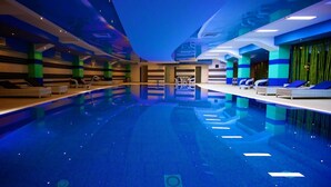En moderne indendørs swimmingpool i blå farve, omgivet af liggestole og et reception område.