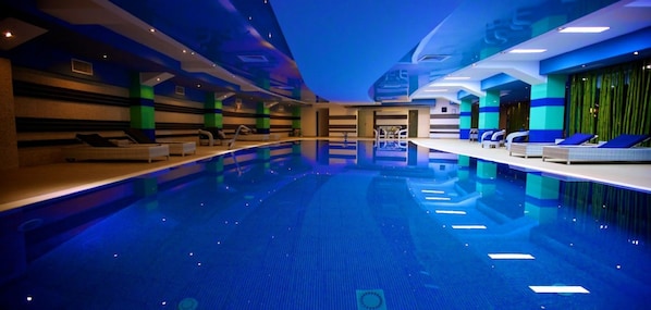A modern indoor swimming pool with a blue hue, surrounded by lounge chairs and a reception area.