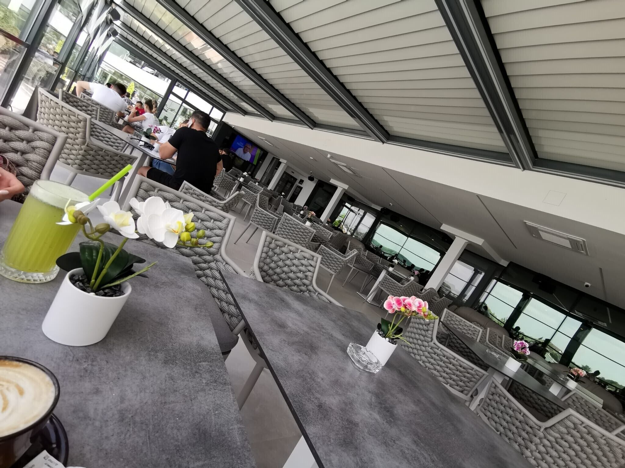 A modern outdoor seating area with cushioned chairs and tables, potted plants, and a large screen displaying a program.