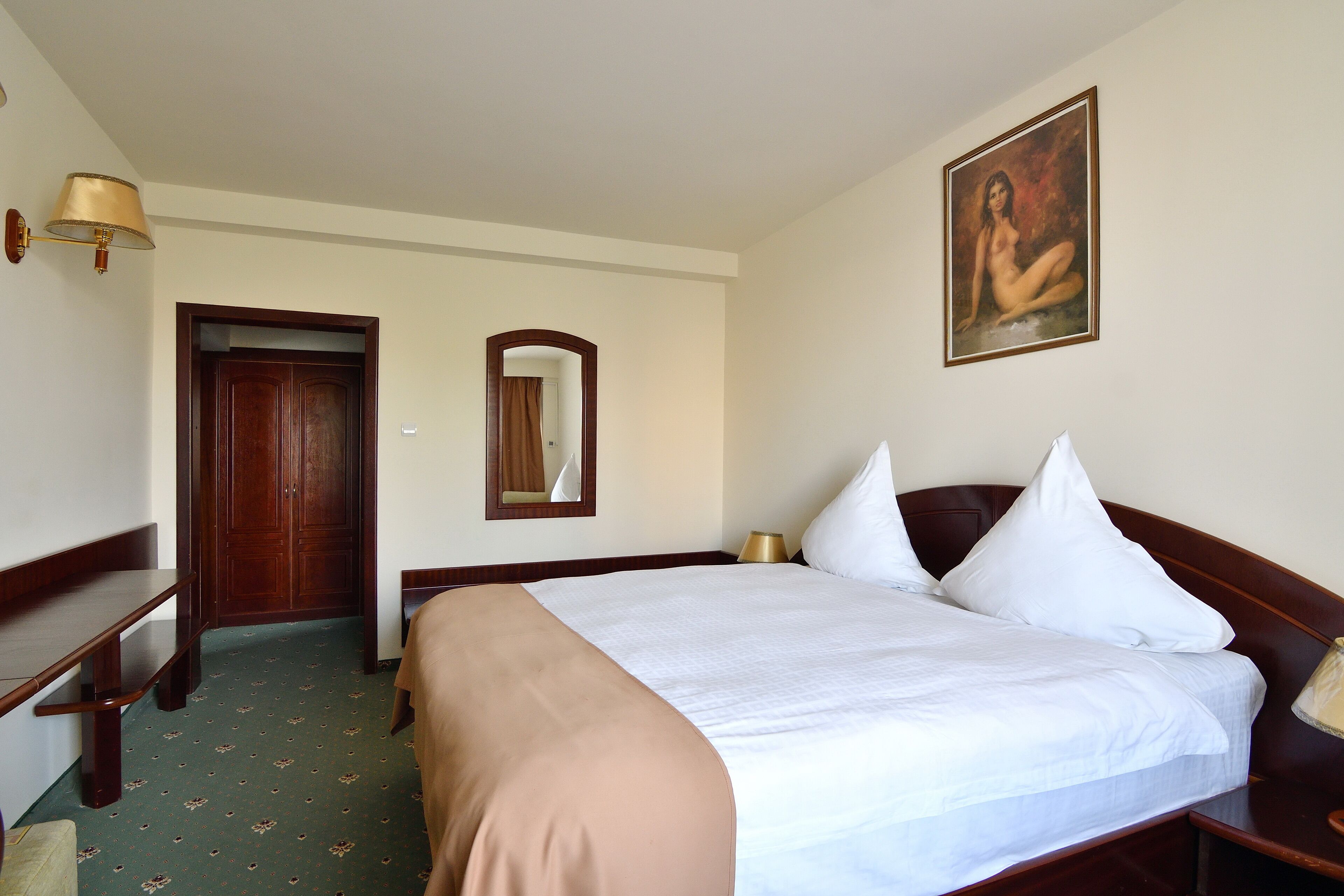A hotel room with a large bed, a wooden desk, a mirror, and a painting on the wall.